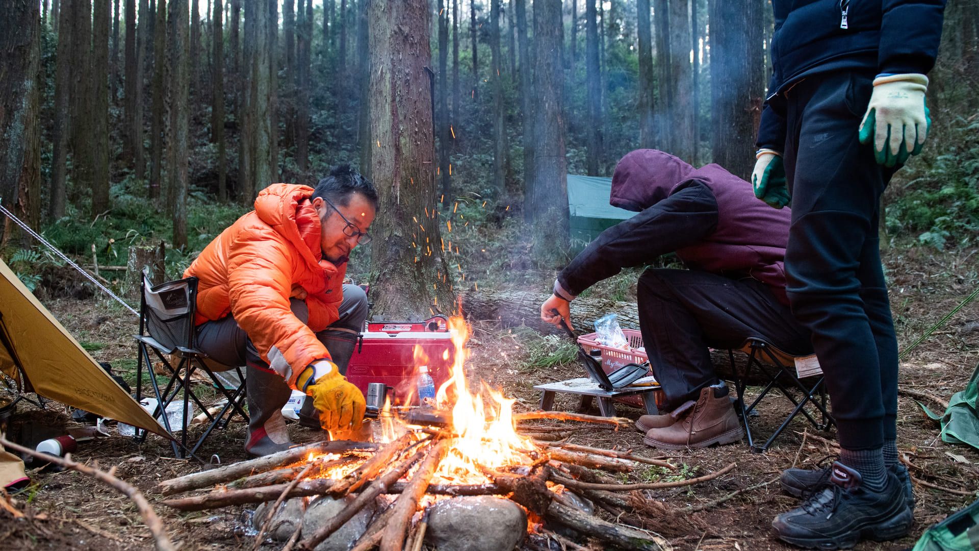 夕暮れの杉林で、焚き火に手をかざしながら食事の支度をする人たち